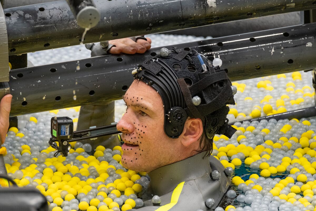 Sam Worthington wears marker dots on his face while in the water, surrounded by floating yellow and gray ball-like devices.