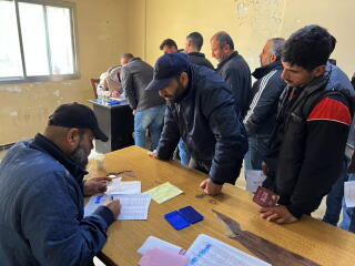 Two men standing at a desk loom over a man filling out paperwork, with other people at another desk