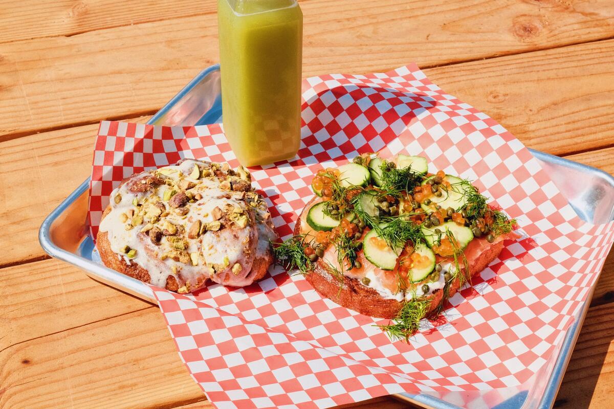 Gravlax-and-ikura smørrebrød on red-and-white checkered paper with a cardamom bun and green juice on a wood table at Piknik