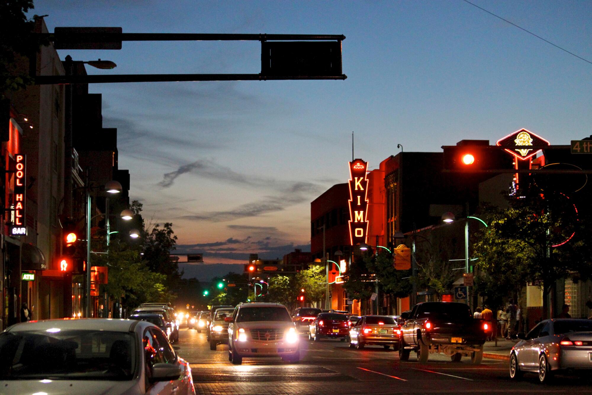 Cars drive down a city street lined with buildings and neon signs at twilight