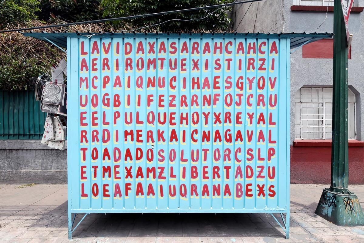 A blue magazine kiosk is covered in a grid of hand-painted letters that reveals phrases like "La Vida" and "El Pulque"