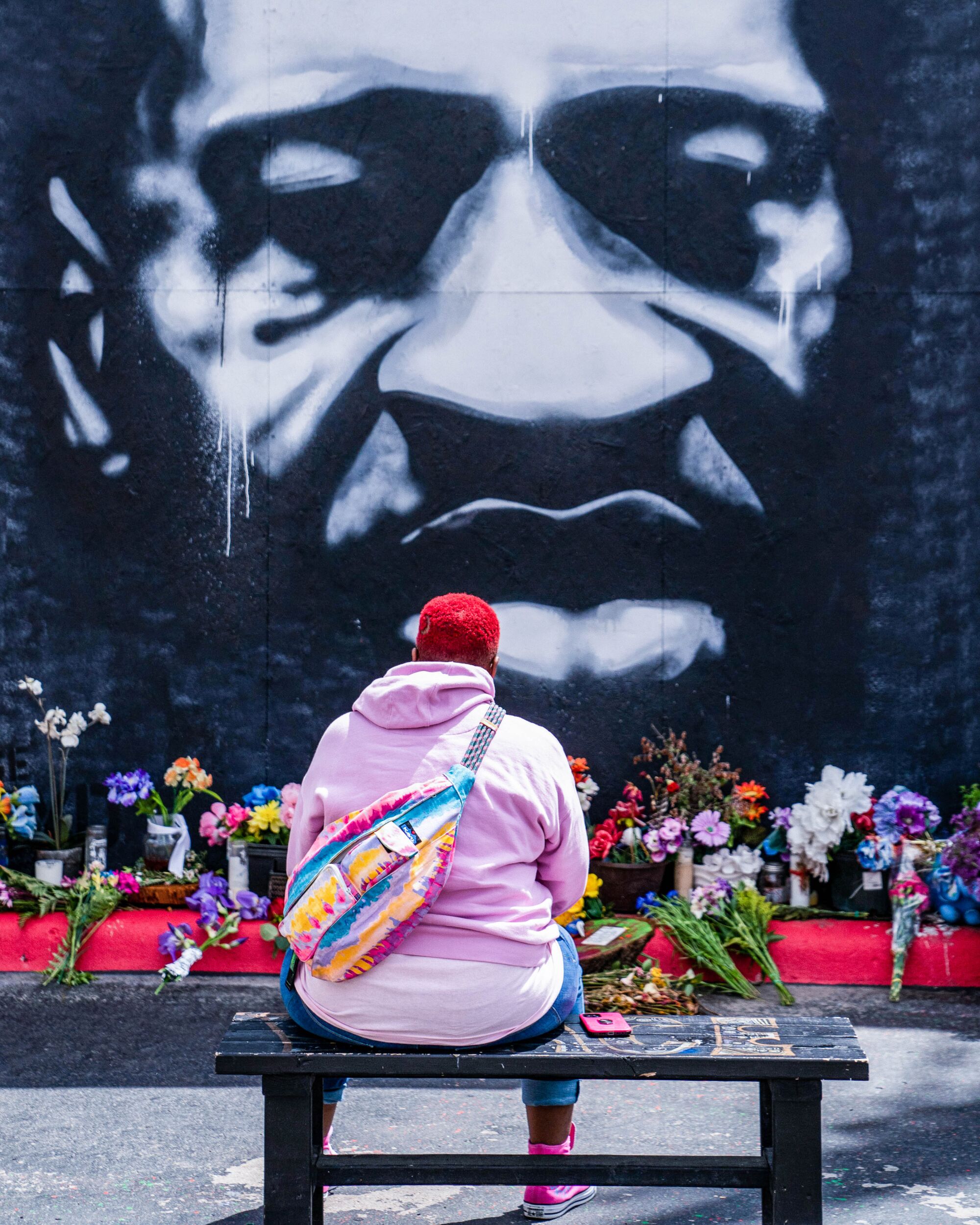 A visitor in a pink hoodie and pink hair sits in front of a mural of George Floyd. Below it is a row of flowers.