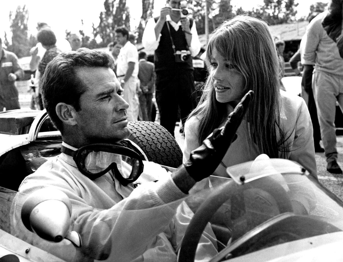 James Garner sits in a racing car, wearing driving gloves and glasses around his neck while Françoise Hardy appears alongside