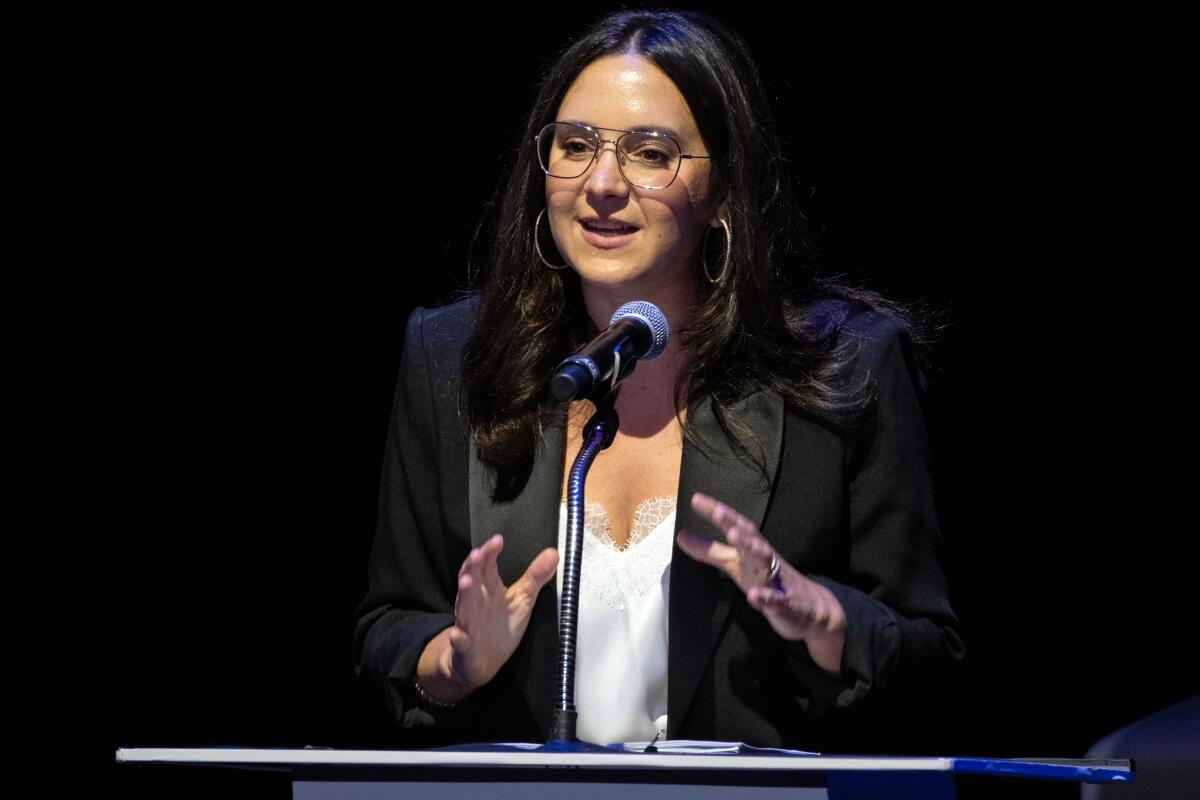 A woman in glasses gestures while speaking at a podium.