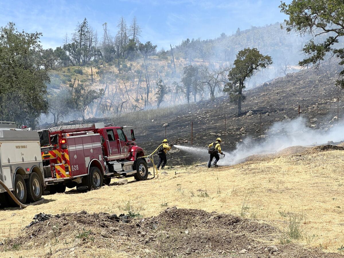 Two firefighters, one aiming a hose from the truck nearby, battling flames on a charred hillside
