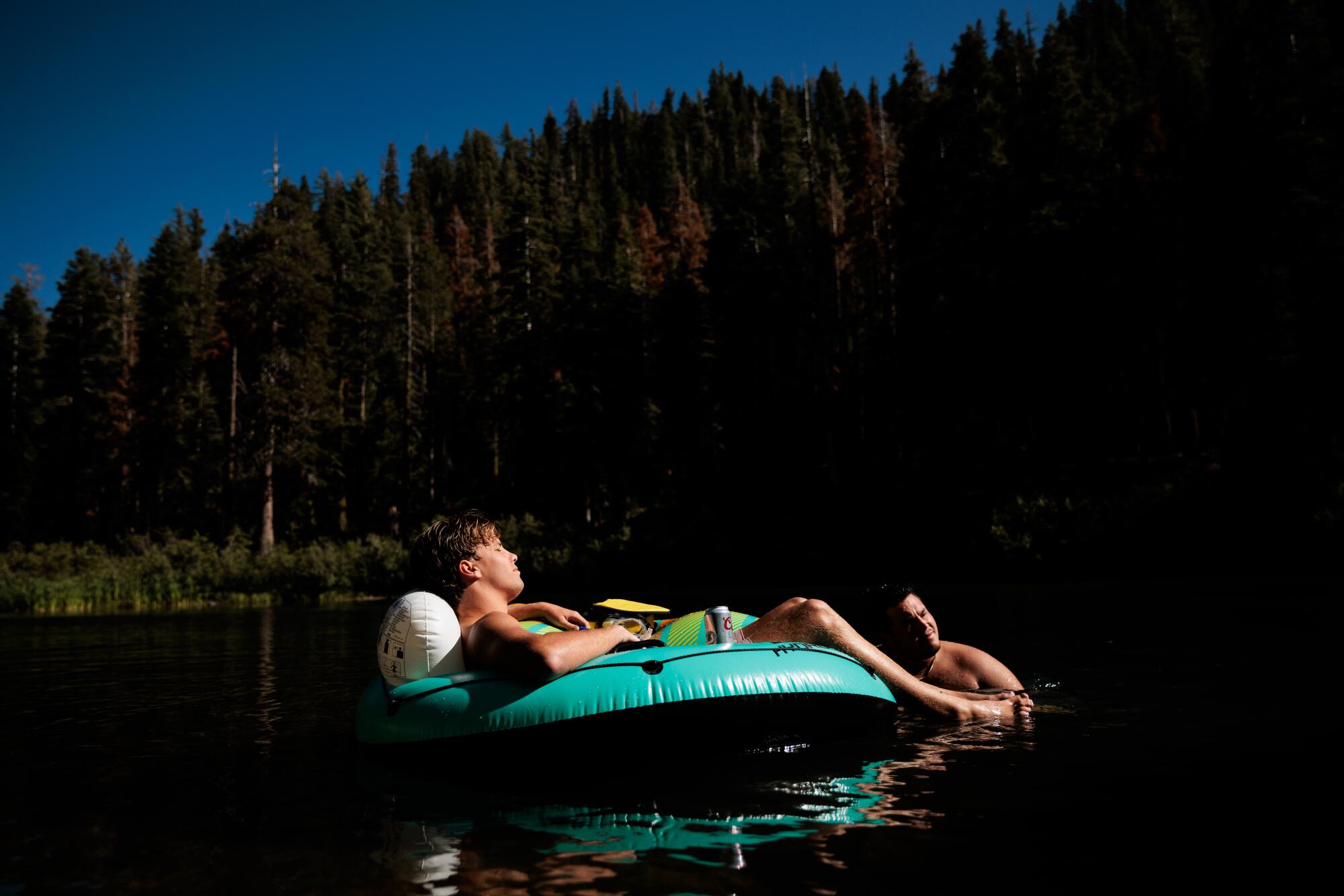 Two people with an inner tube on a lake ringed by conifers.