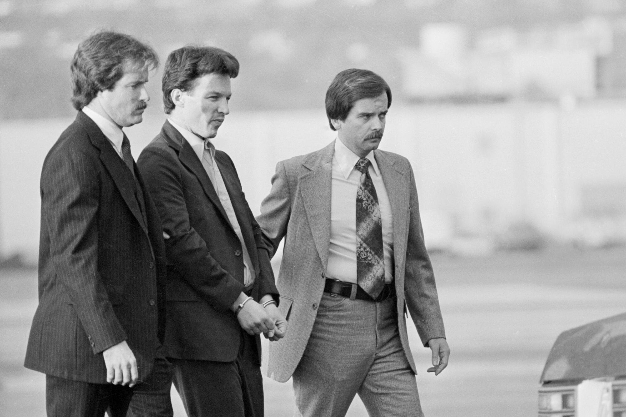 A black-and-white photo of a man in a dark suit and handcuffs, flanked by two men in suits and ties.