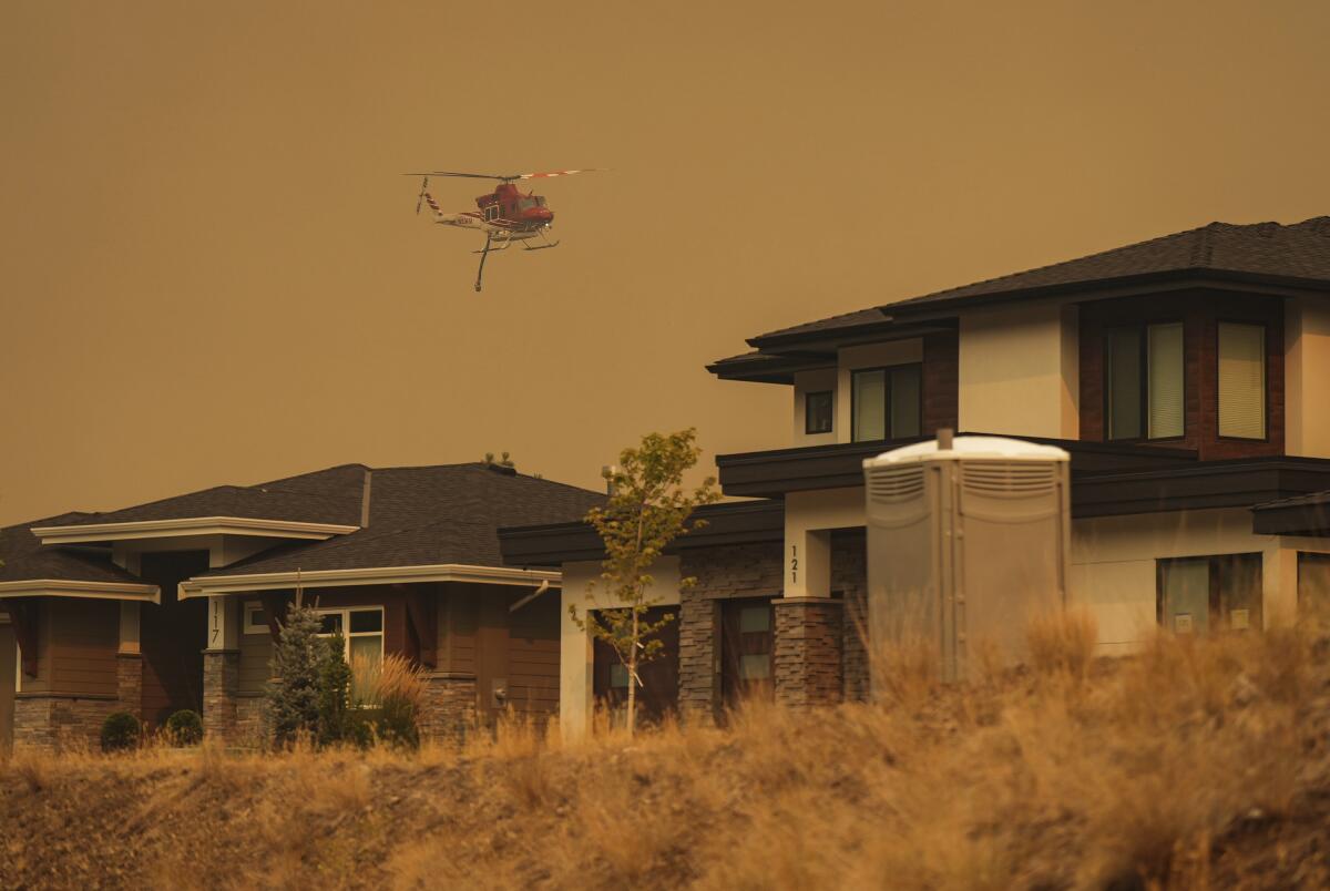 In smoky brownish-orange air, a firefighting helicopter flies over houses in Kelowna, British Columbia