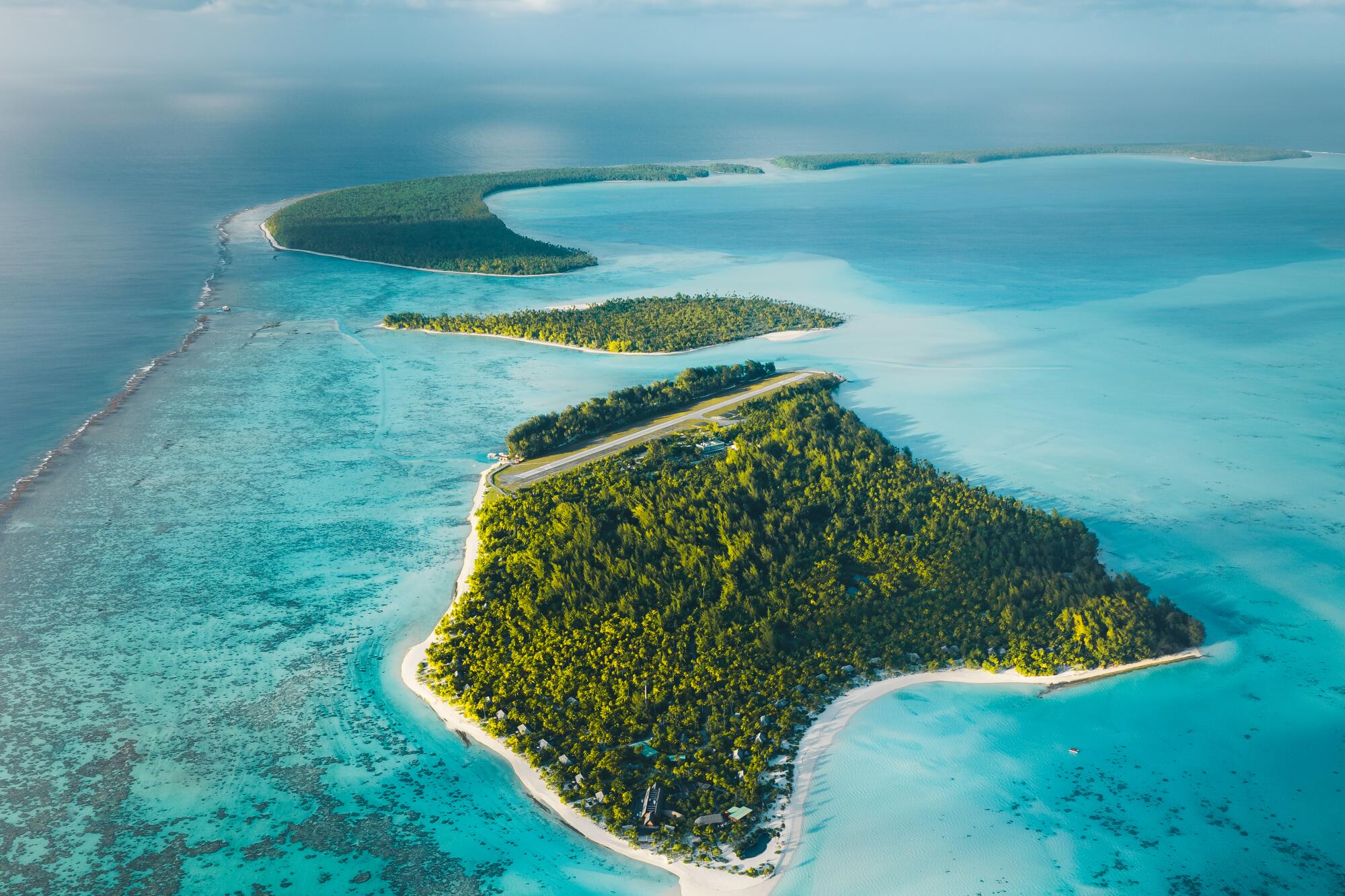 An atoll covered in greenery, in turquoise ocean waters under a sky with puffy white clouds