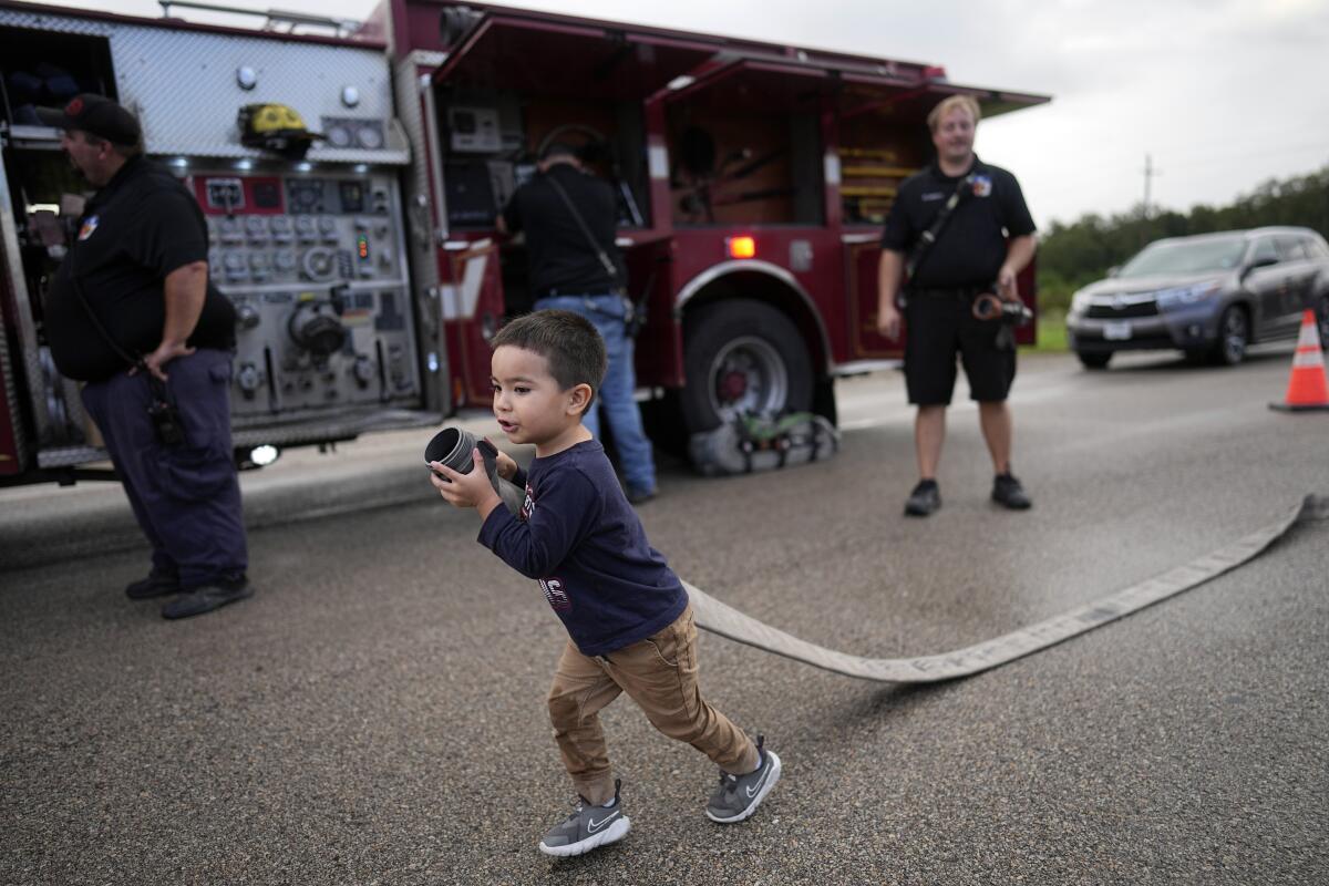 A small boy carrying a fire hose down a street next to a firetruck as adults stand by