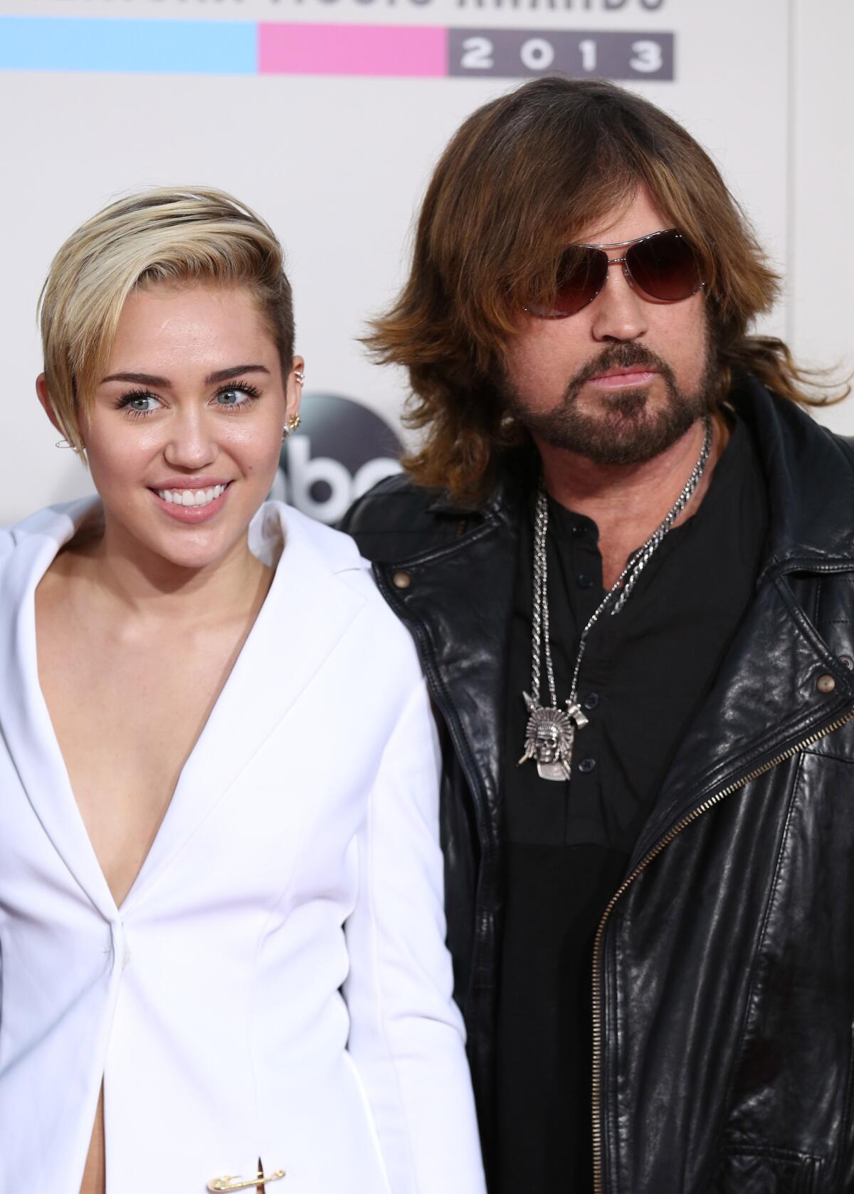 Miley Cyrus with short blond hair and a white suit jacket and Billy Ray Cyrus wearing all black pose in front of backdrop