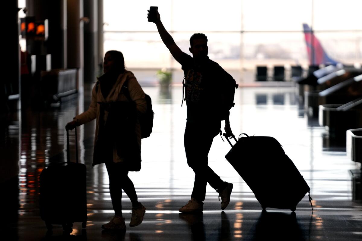 Two silhouetted figures walk across a backlit airport hall pulling a suitcase behind.