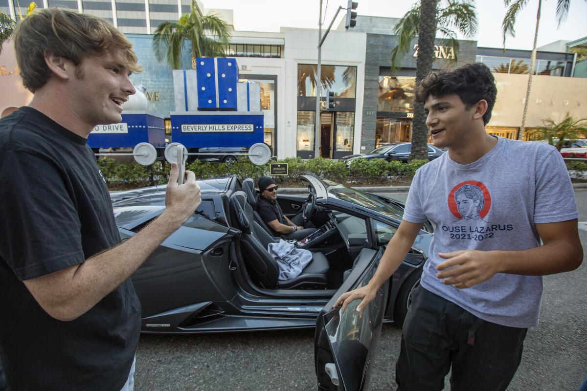 A man stands next to a sports car holding up his phone to record another man 