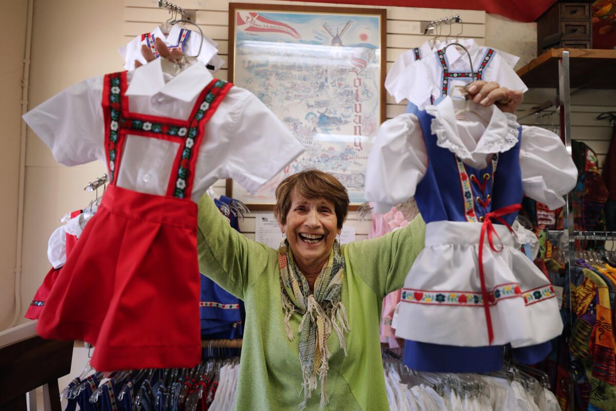 A laughing woman inside a shop holds up a child-size traditional Danish outfit in each hand.
