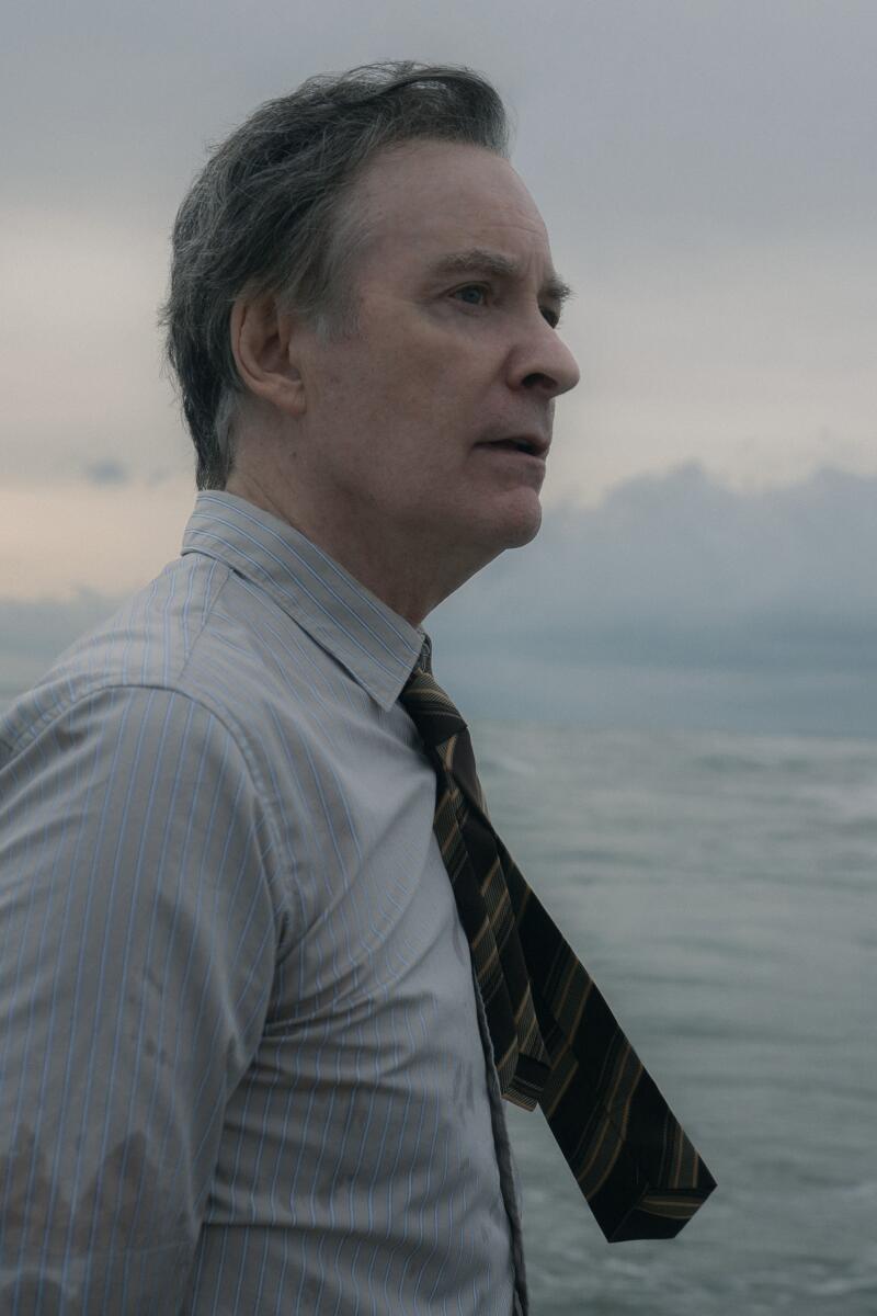 A man in a shirt and tie, seen from the waist up, stands with his back to the ocean.