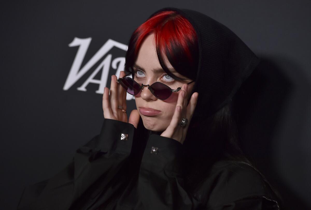 Billie Eilish with red ombre hair, holding down her sunglasses with her hands at a red carpet against a black background