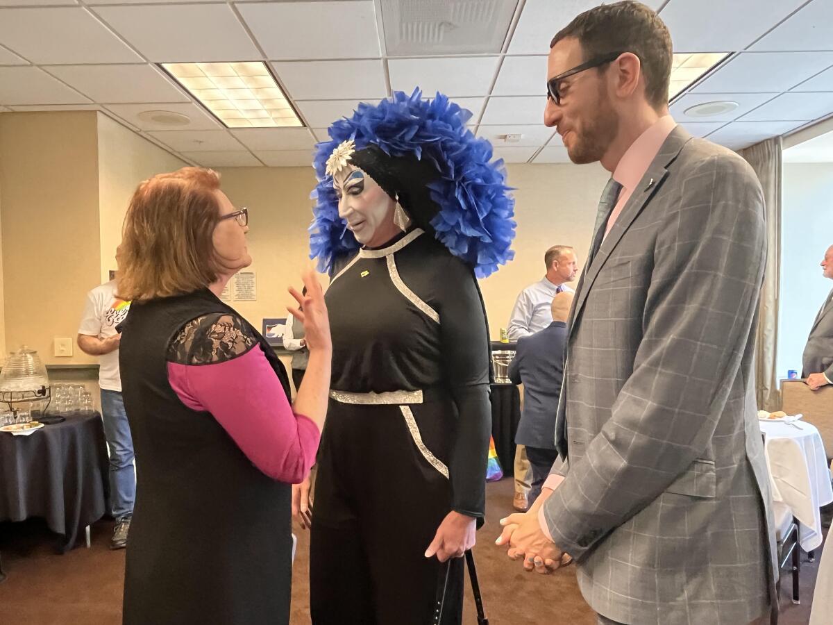 A drag activist, wearing a feather boa headdress, center, talks to two lawmakers, one female, left, and male, right. 