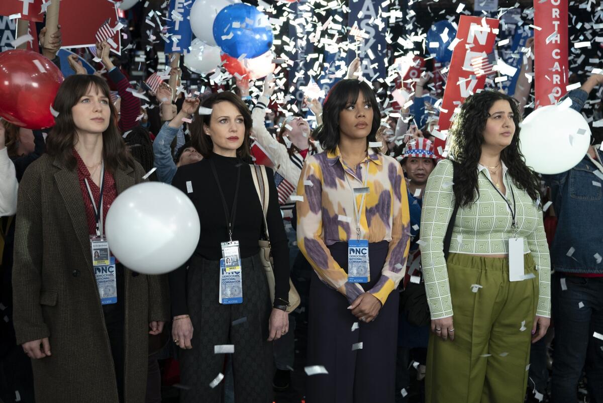 Four women wearing press badges look to their left as balloons and confetti fall from the ceiling.