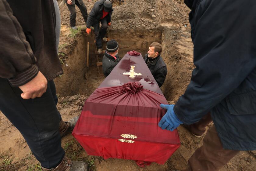 Two men stand in a large hole and hold a red casket being lowered into the grave. 