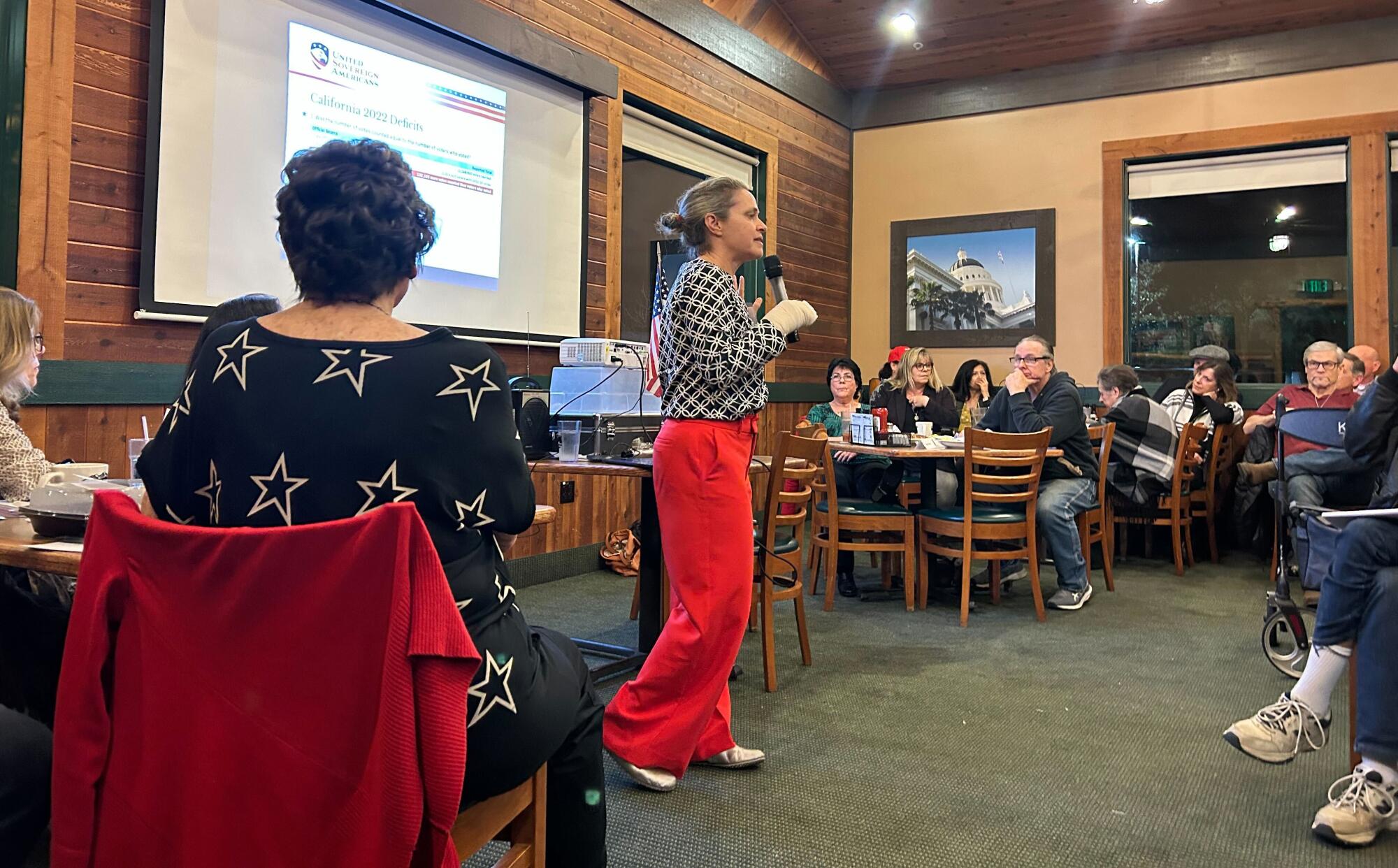 A woman in a printed top and red pants stands as she addresses people seated at tables in a restaurant 