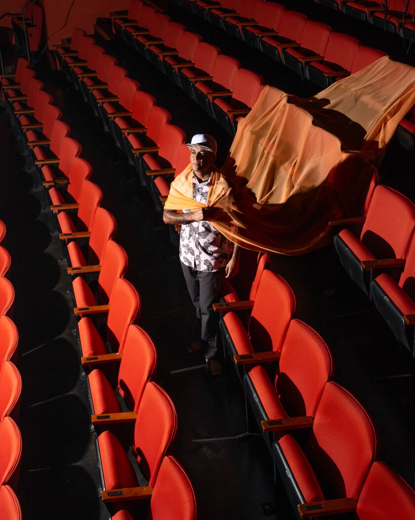 D'Lo in an empty theater with a swath of silky fabric trailing over two rows of seats behind him.