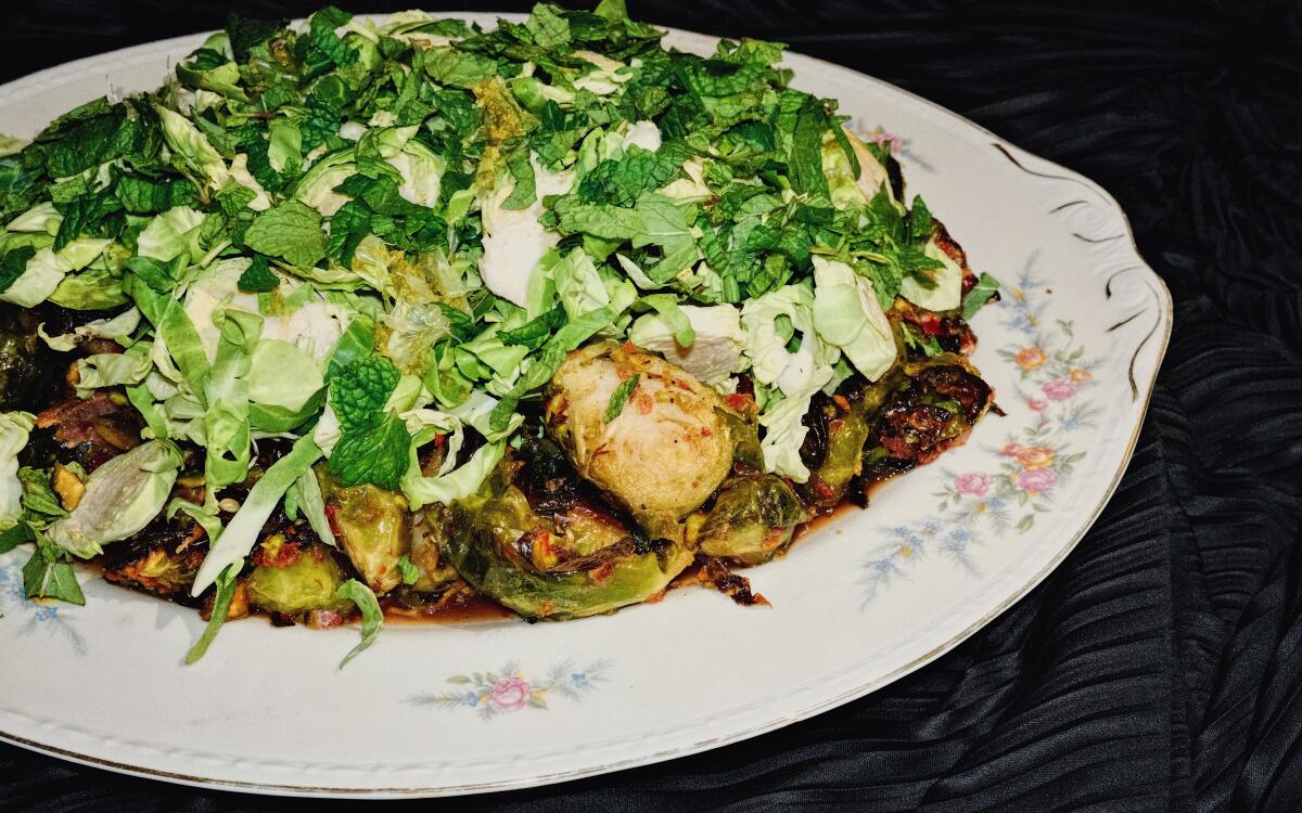 A floral porcelain platter of Brussels sprouts cooked and raw, with bacon, mint and pistachio, on a black cloth background
