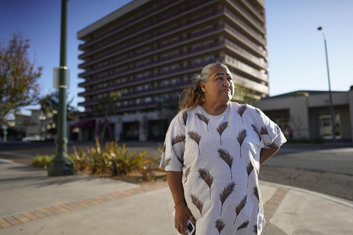 A woman with a graying ponytail stands on a sidewalk in front of a mid-rise hotel building.