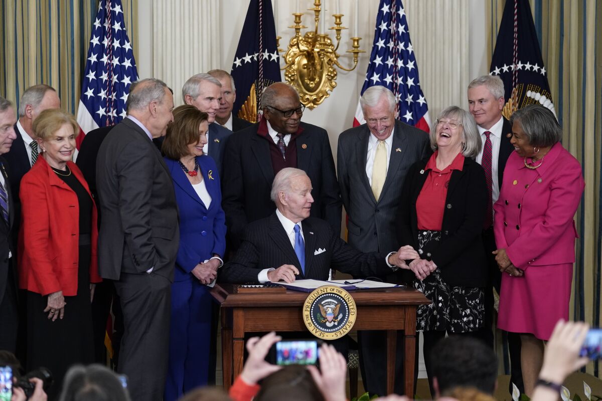 President Biden sits at a desk, signing a bill, surrounded by lawmakers 