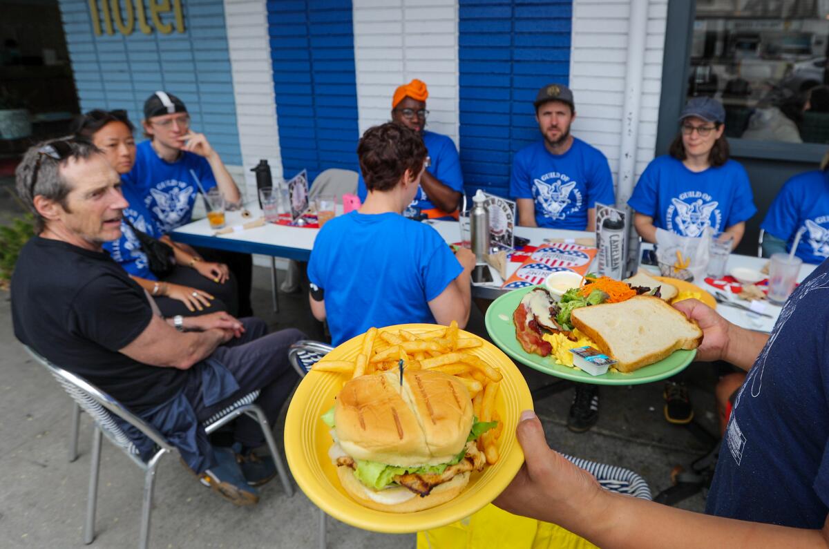 WGA members clad in blue guild shirts sit at a patio table at Swingers Diner. In the foreground a server carries burgers.