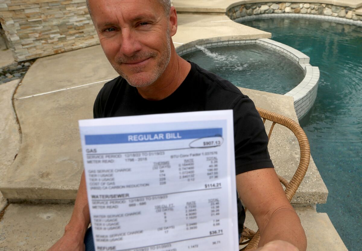 A man wearing a black T-shirt sits in front of a hot tub and swimming pool, holding a utility bill.