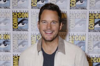Chris Pratt in a cream-colored jacket and a black shirt smiling and standing in front of a Comic-con backdrop