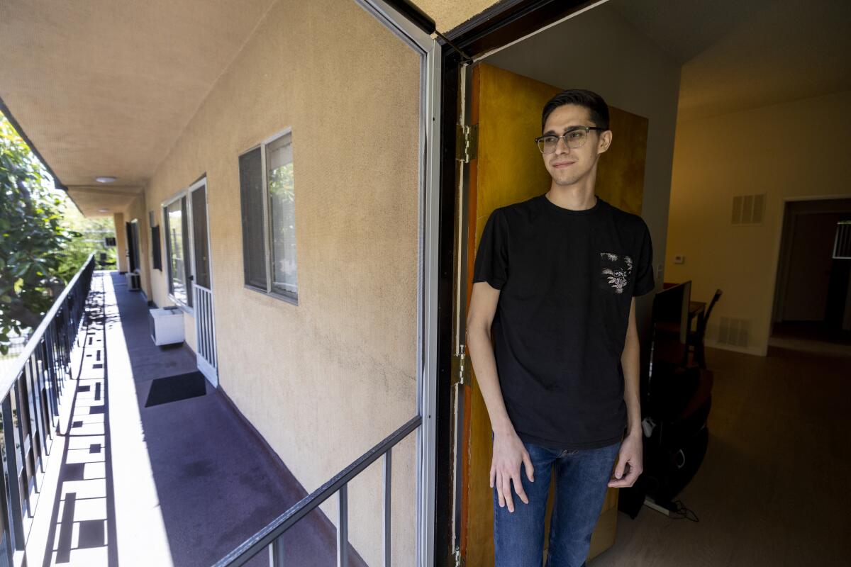 A young man stands inside the open front door of an apartment.