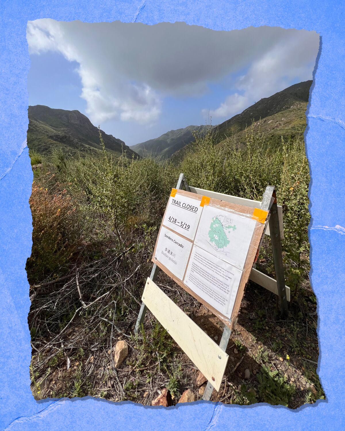A sign that says "Trail Closed" sits in a grassy area with mountains and blue sky in the background.