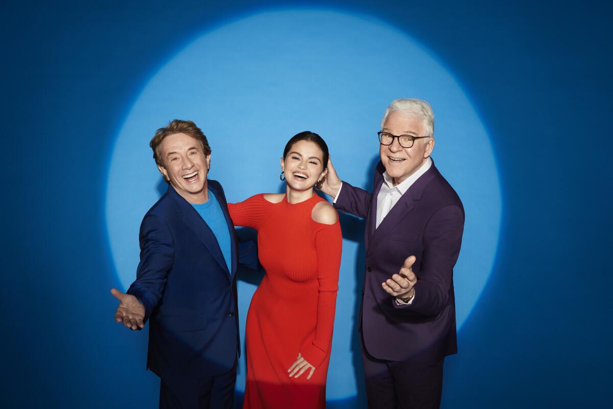 Two men flank a woman in red before a blue backdrop in a spotlight.