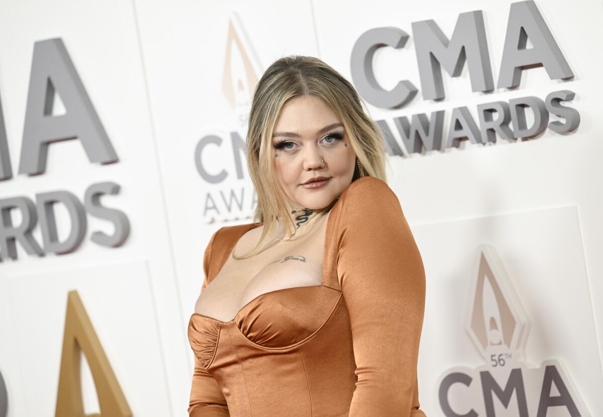 Elle King raises her left shoulder to her chin while posing in a bronze dress at an awards show