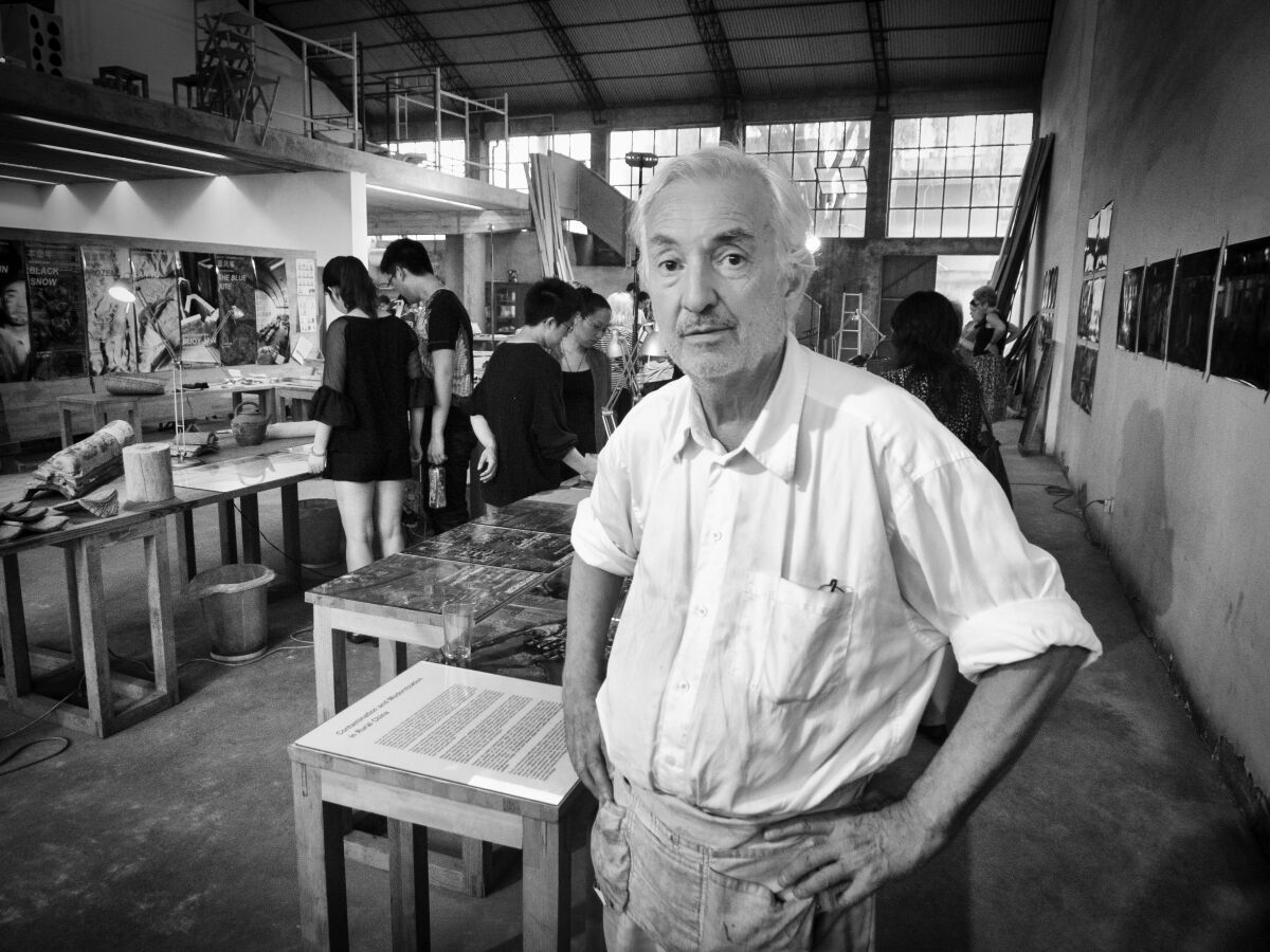 A gray-haired man in a white shirt and workshop apron stands with his hands on his hips inside an architectural studio