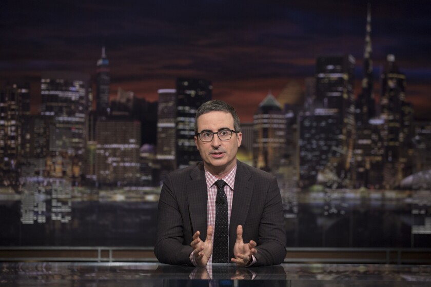 John Oliver sitting behind a desk with the New York skyline behind him.