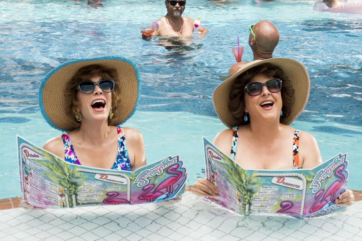 Two women in sunglasses and floppy hats hold menus at a swim-up bar