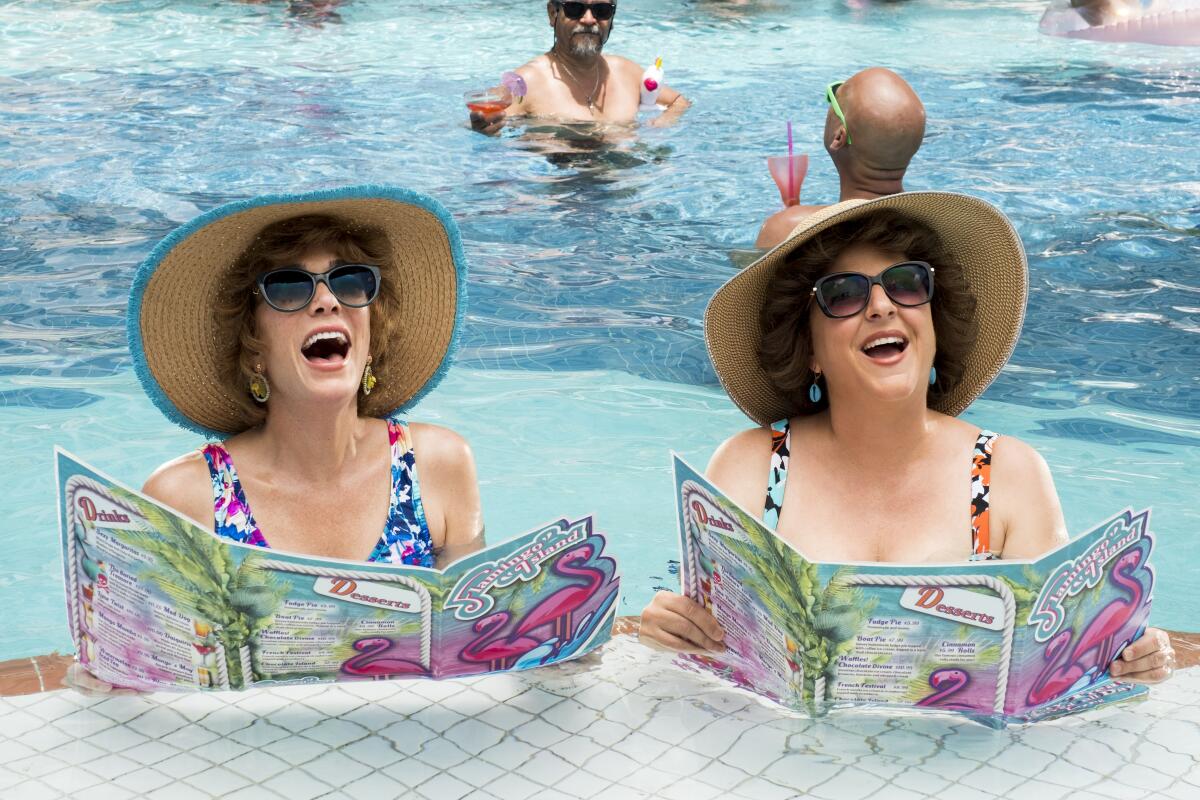 Two women stand in a swimming pool wearing sunhats and holding colorful menus.