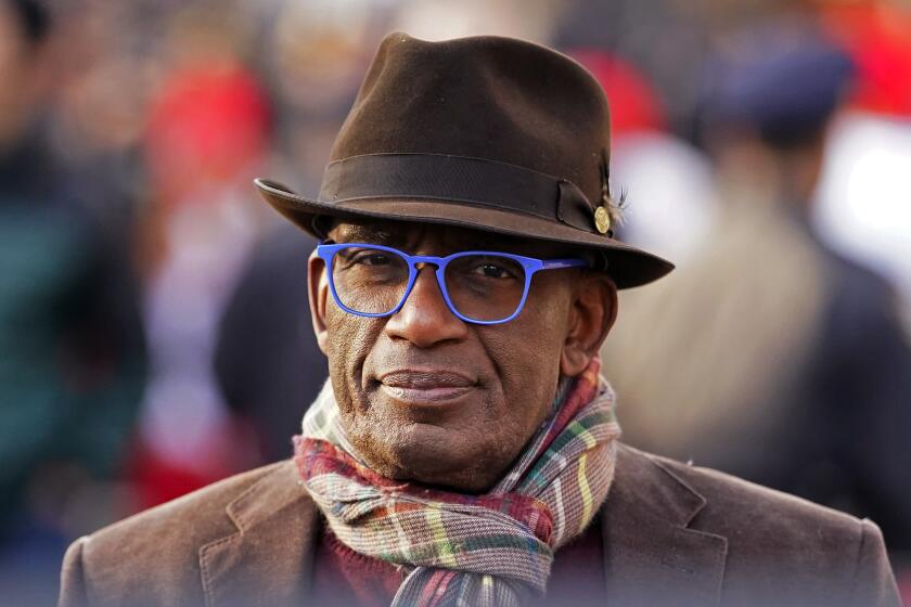 A man staring into the camera while wearing a colorful scarf, blue-framed glasses and a brown fedora