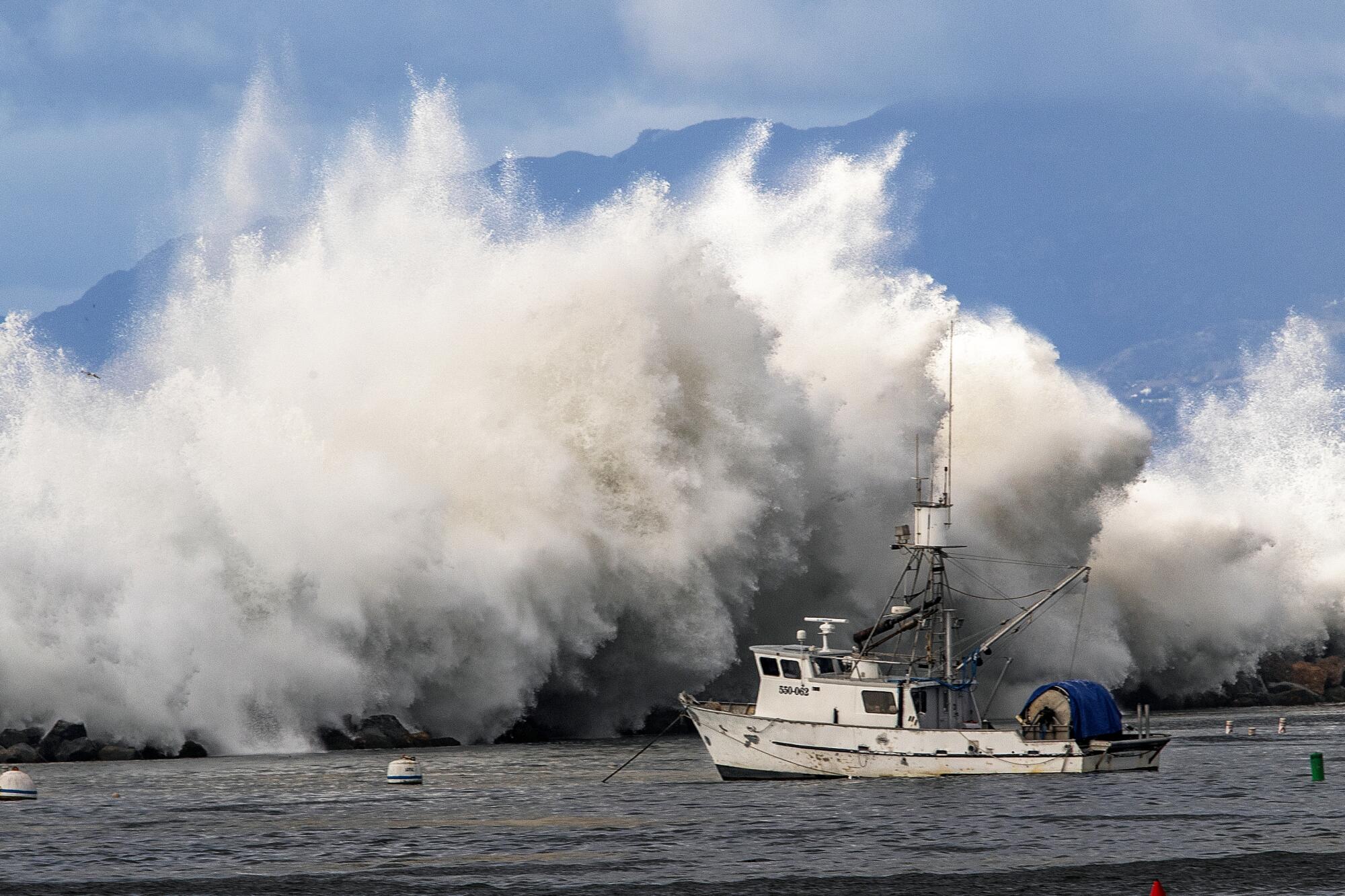 A giant wave crashes over a jetty as a fishing boat floats in the foreground.