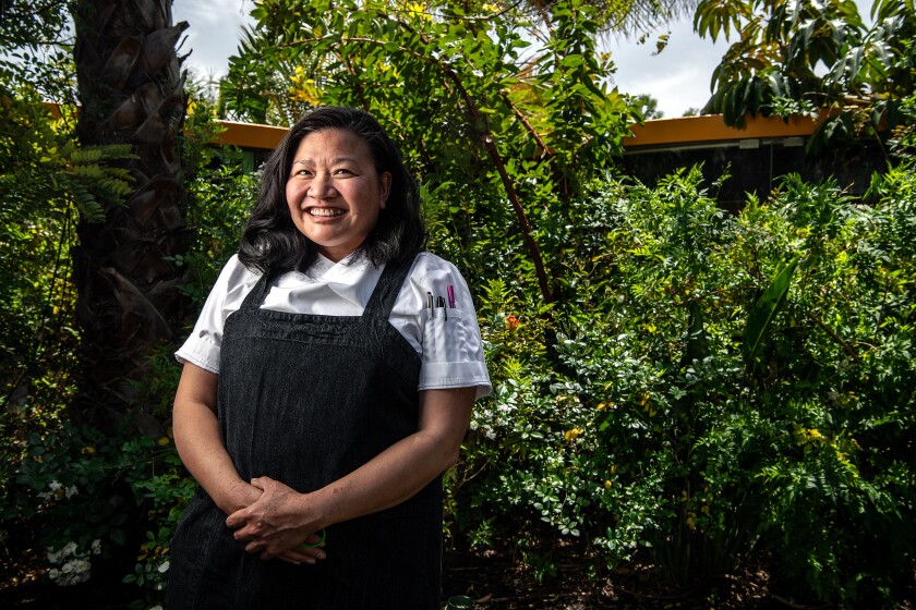 A woman smiles and clasps her hands in front of her as she stands in front of greenery.