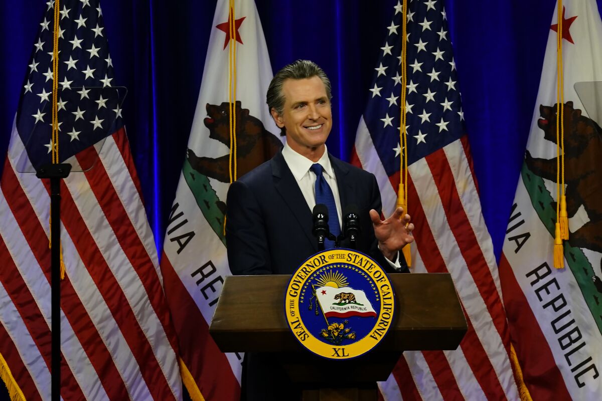 A man in a dark suit and blue tie smiles as he speaks at a lectern with the California governor's seal, in front of flags