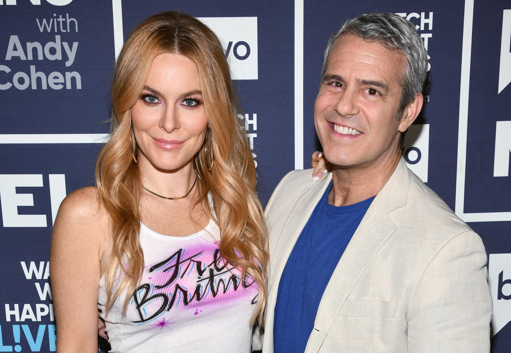 A woman with wavy blond hair in a white tank top stands next to Andy Cohen in a beige suit jacket and blue shirt