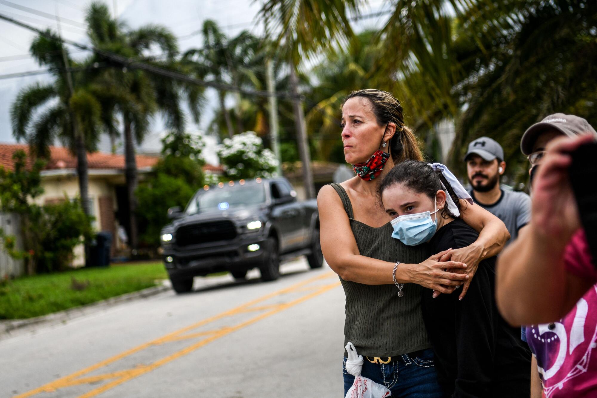 A woman, standing with others along a street with palm trees, hugs her daughter. 