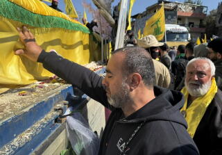 A man touches a casket draped with yellow cloth with green trim as people stand nearby