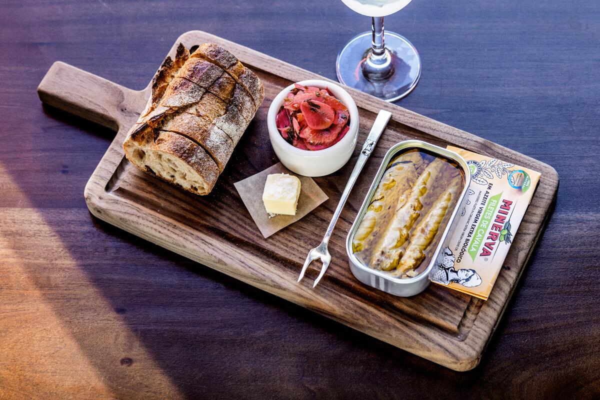 Mackerel in a tin on a board with a sliced baguette and pickled vegetables.