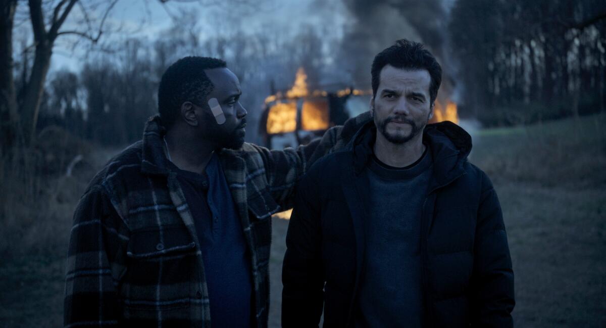 A man in a flannel shirt with a bandage on his cheek puts an arm around a man as they walk away from a burning van.