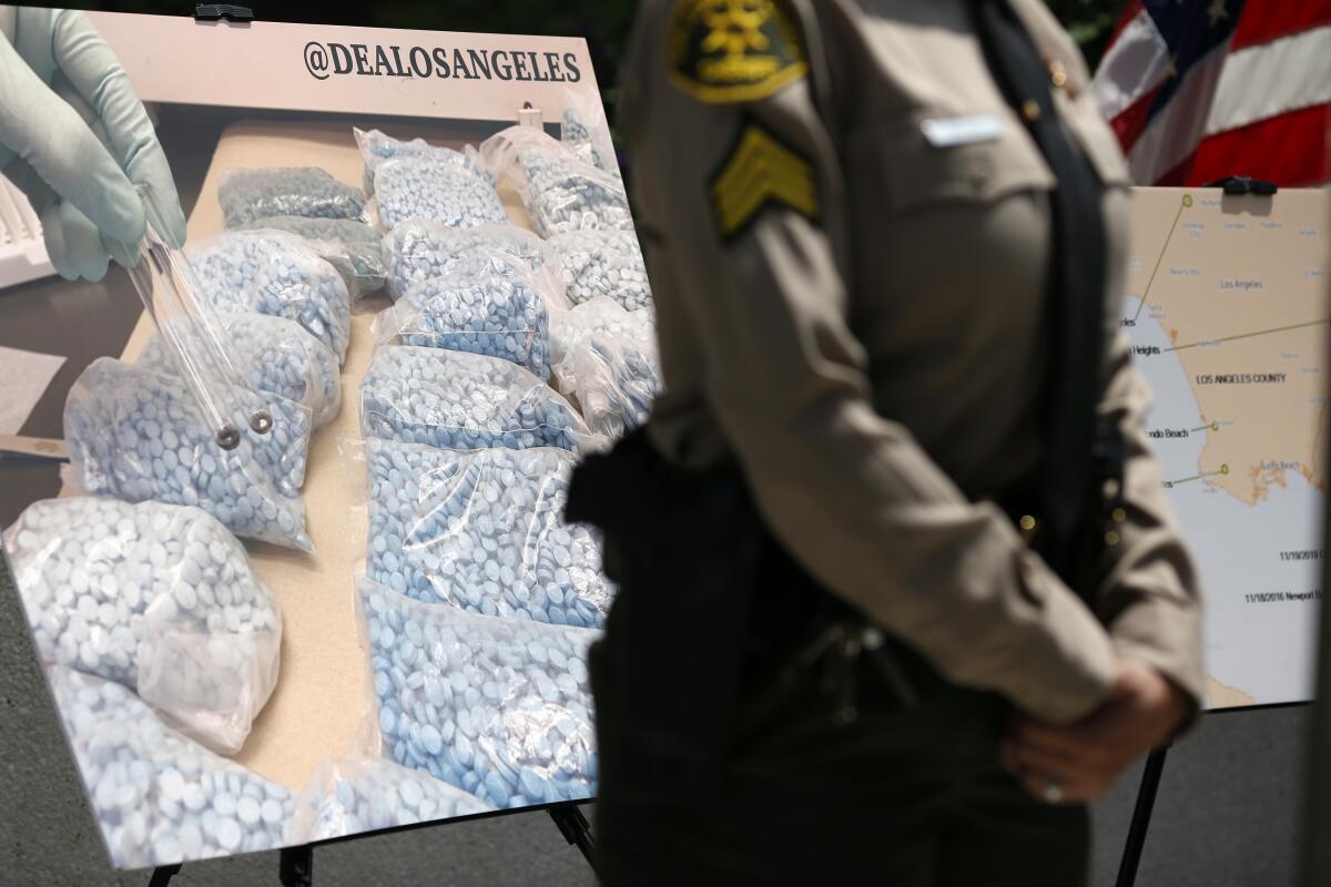 Law enforcement officer stands in front of an enlarged photograph of bags of counterfeit fentanyl pills at a news conference