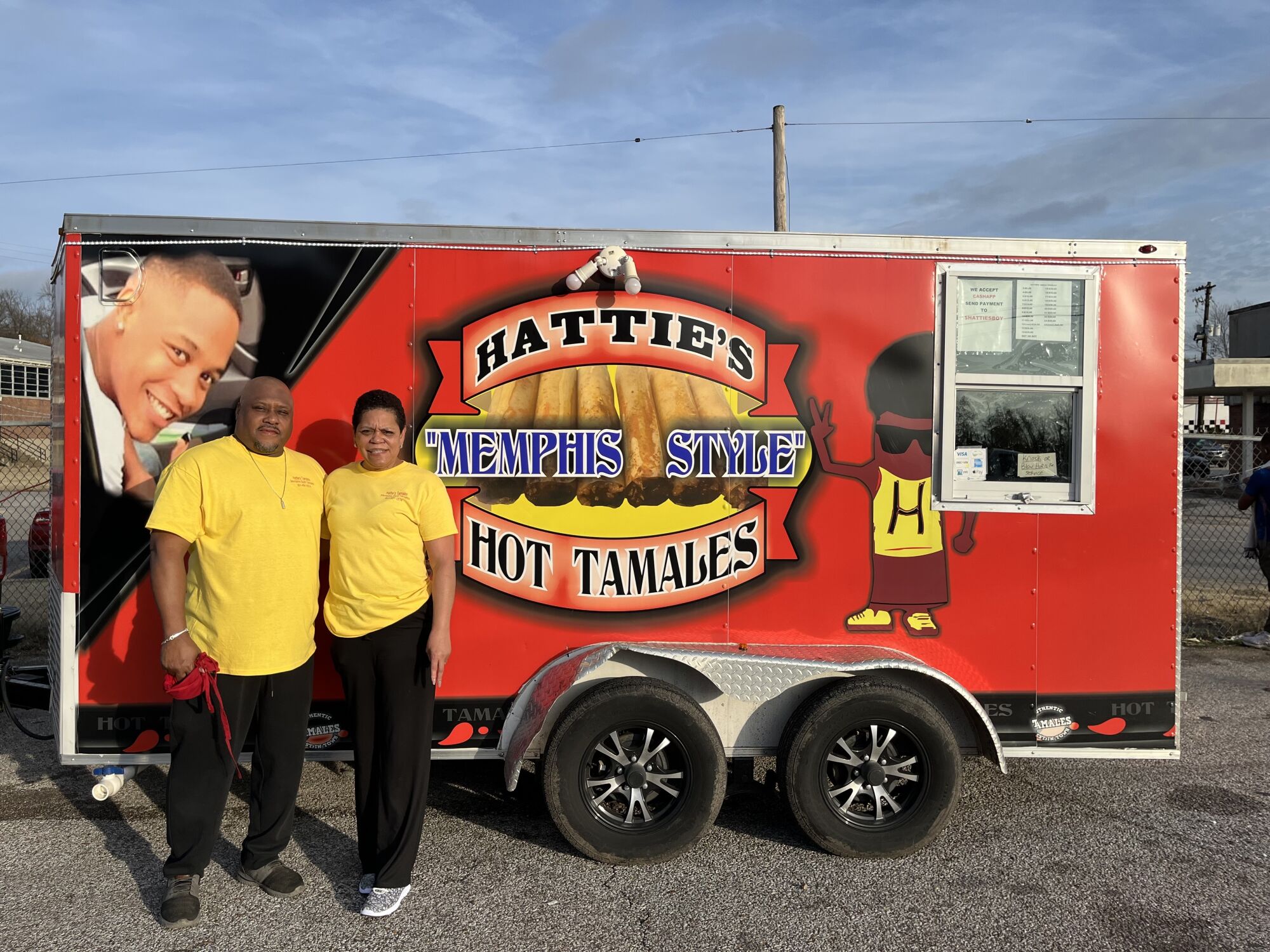 A man and a woman in yellow shirts stand outside their trailer, which says Hattie’s Tamales.
